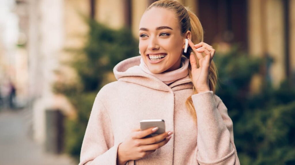 Why does the FTSE 100 keep outperforming the S&P 500? Smiling white woman holding iPhone with Airpods in ear